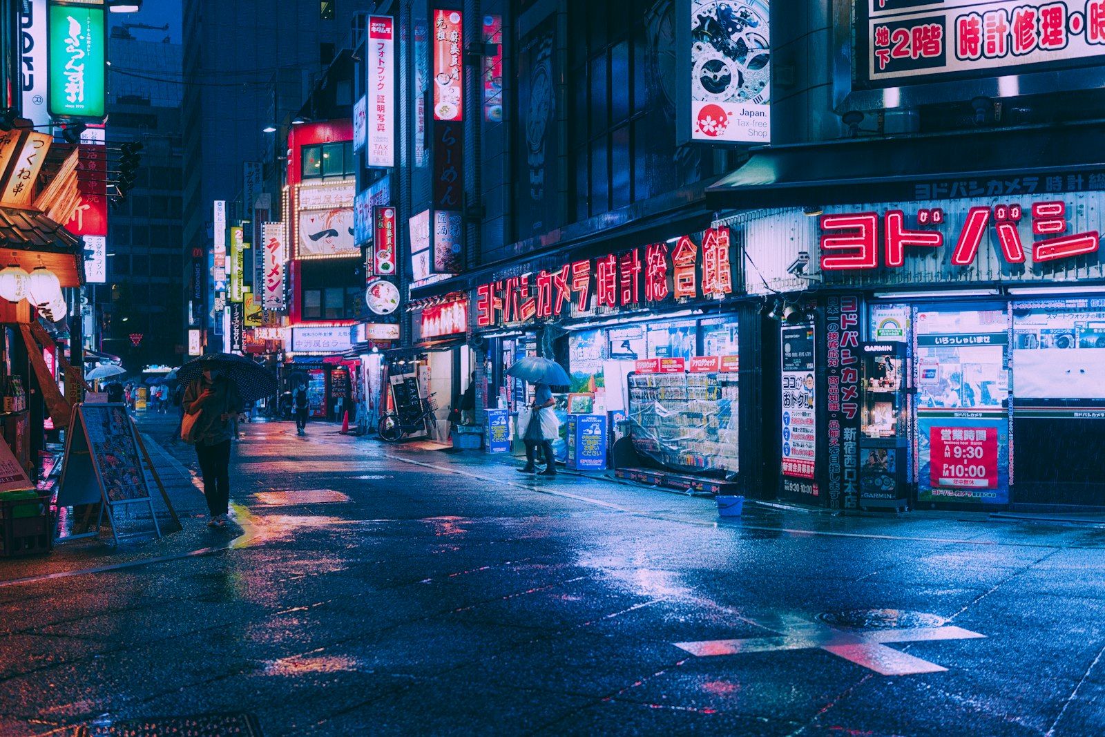 People walking a busy neon-lit Shinjuku street at night, where touts approach tourists outside hostess club buildings