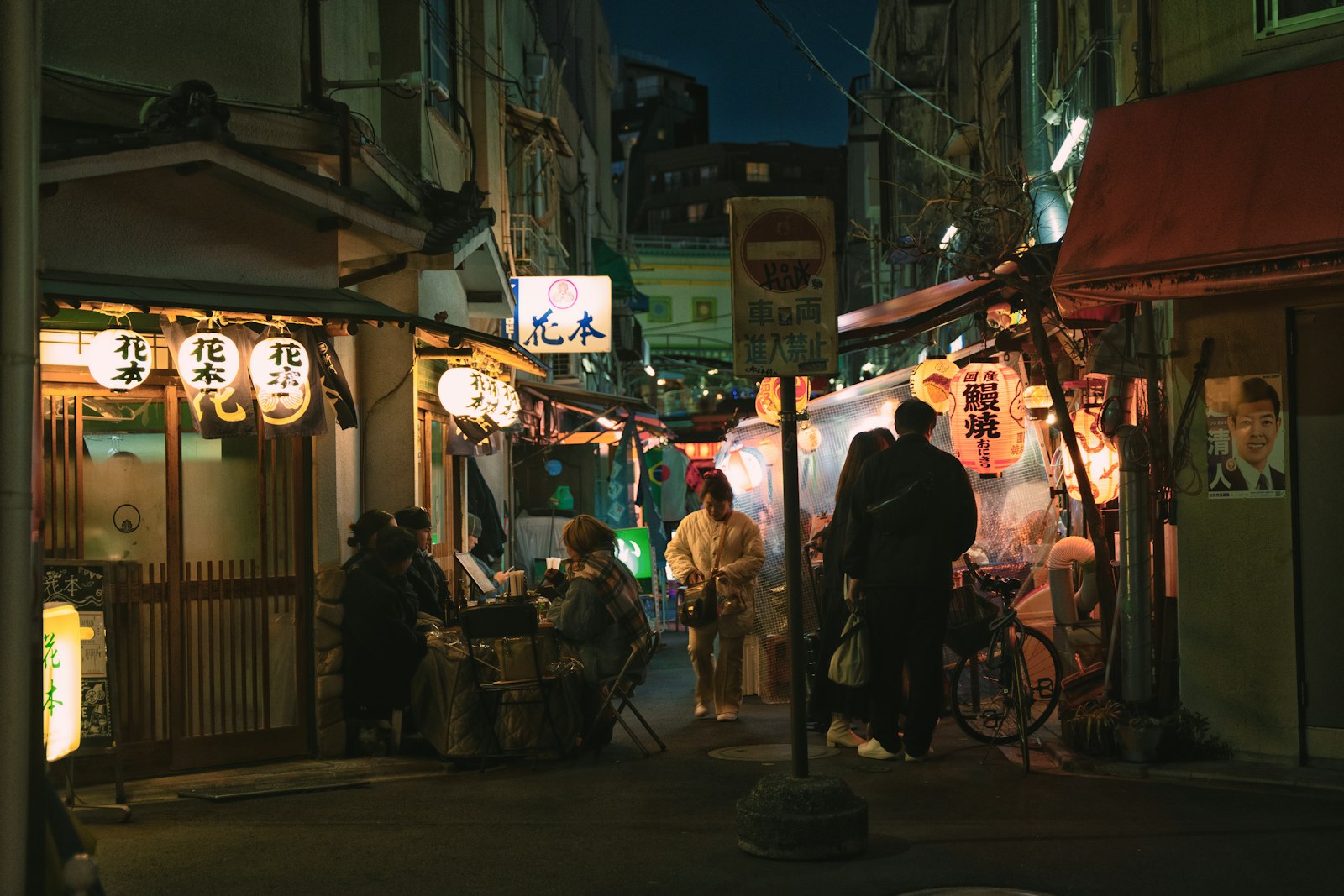 Customers drinking beer shoulder to shoulder at a small izakaya counter in a Tokyo alley